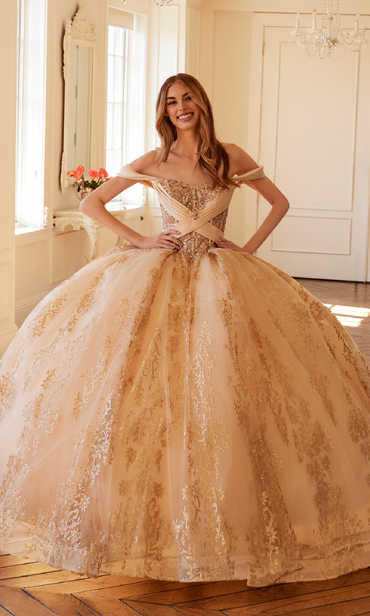 A woman stands indoors wearing the Quinceanera Dress JT1488H By Juliet, a champagne gown with gold embroidery, a corset bodice, and a full ball gown skirt.
